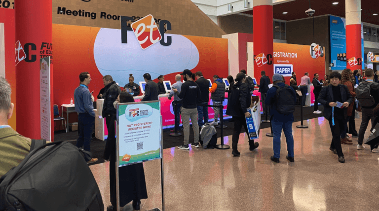 Attendees line up at registration kiosks for the FETC conference in a large convention hall with red pillars and event signage. Some people check in at kiosks while others walk or wait nearby.